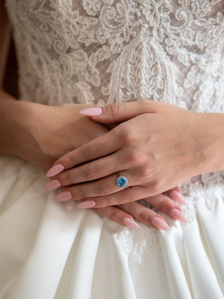 A bride’s hands with a blue gemstone ring resting on her lace wedding dress.