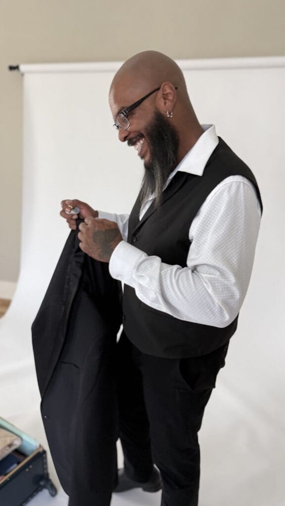 A bearded man in a white dress shirt and black vest laughs while holding a suit jacket.