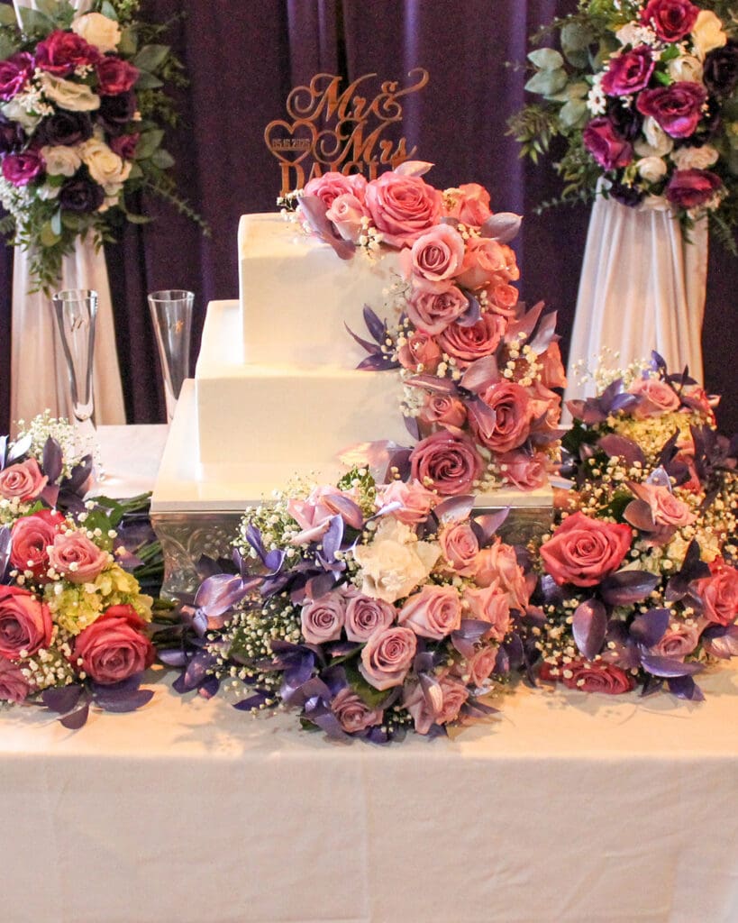 Three-tiered white wedding cake with custom clothing detailing, a Mr. & Mrs. topper, and pink and purple floral bouquets.