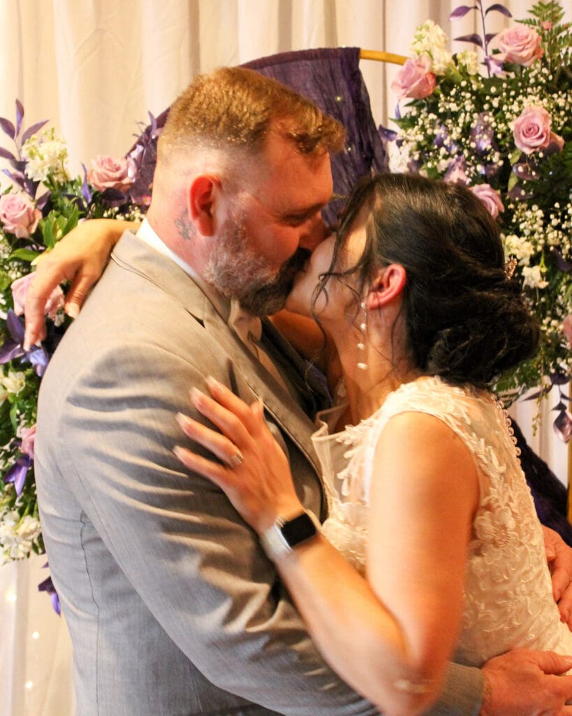 Bride and groom in custom wedding attire kissing before floral arrangements.