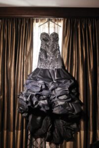 Strapless black gown with lace bodice and ruffled satin skirt hanging before brown curtains and a sheer lace-covered window.