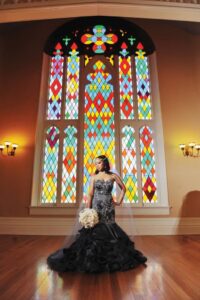 Woman in a black strapless gown holds a bouquet before a colorful stained glass window in an elegant, bright room.