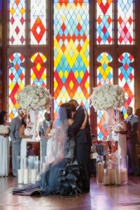 Bride and groom embrace at the altar with their wedding party, colorful stained glass, flowers, and candles in the background.