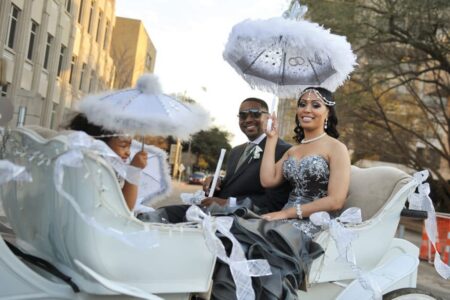 A formally dressed couple and a child ride in a white carriage with decorated umbrellas, buildings and trees behind them.