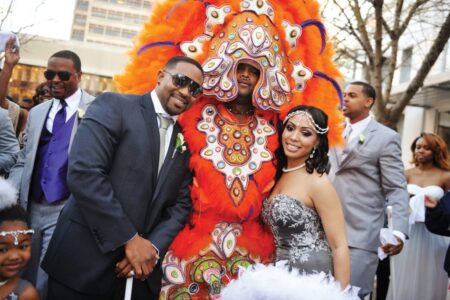 Wedding couple poses with a person in a bright orange feathered costume outdoors, smiling guests in formal wear behind them.