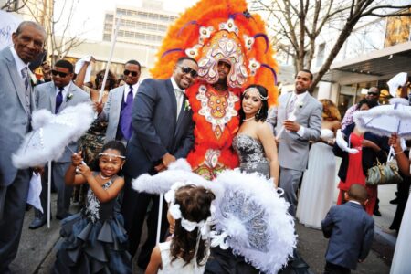 Wedding party outdoors with feathered umbrellas; guests in formal attire and vibrant costumes smile and celebrate.