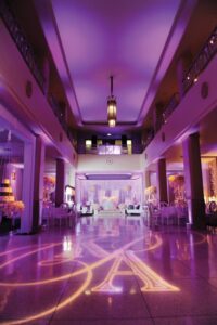 Ballroom with purple lighting, elegant tables, clear chairs, floral centerpieces, and a monogram on the shiny floor.