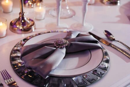 Jeweled gray napkin on a decorative plate with candles, cutlery, and wine glasses atop a white tablecloth.