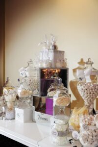 Elegant glass jars with white candies and treats on a table with white ribbons, centered by a mirrored “Then Now and Forever” sign.