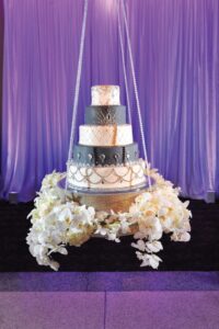 Five-tiered hanging cake with white and dark gray layers, crystals, silver accents, and white flowers on purple draped backdrop.