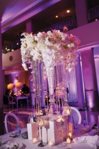 Elegant glass vase centerpiece with cascading white flowers, crystals, and candles on a formal event table under purple lighting.