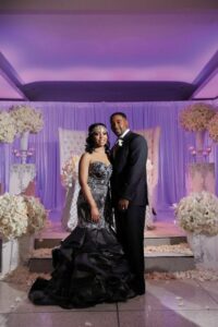 Couple at wedding in formal attire, woman in black gown with silver details, man in tuxedo; white flowers and purple lighting.