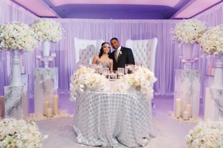 Bride and groom behind a flower-adorned table at a white-and-silver wedding with candles, florals, and draped fabric.