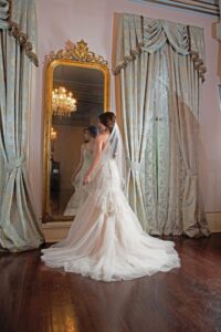 Bride in white gown and veil stands before ornate mirror, reflection visible, with curtains and chandelier behind her.
