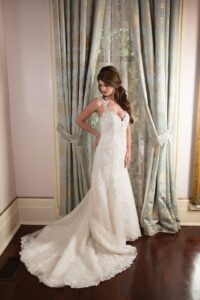 Woman in an elegant white lace wedding dress stands before ornate curtains, looking down with loose waves in her hair.