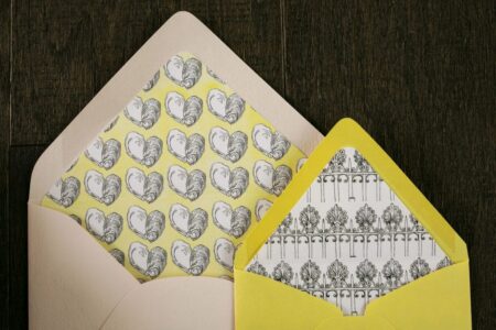 Two open envelopes on dark wood: one cream with a heart liner, one yellow with a black and white decorative liner.