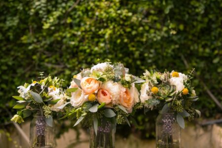 Three glass vases with peach, white, and yellow flowers plus greenery, displayed outdoors against a blurred leafy background.