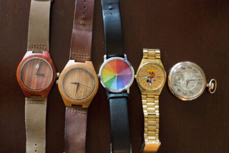 Five wristwatches and a pocket watch with wood, rainbow, cartoon, gold, and vintage designs on a dark surface.