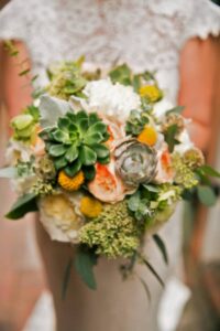 Bride in a lace dress holding a bouquet of succulents, peach roses, billy balls, white flowers, and greenery.