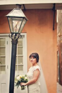 Bride in white dress and veil holding bouquet by vintage streetlamp, in front of peach building with white shutters.