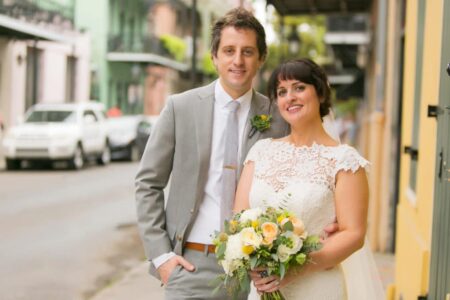 Bride in white lace dress with yellow and white bouquet stands by groom in light gray suit on city street.