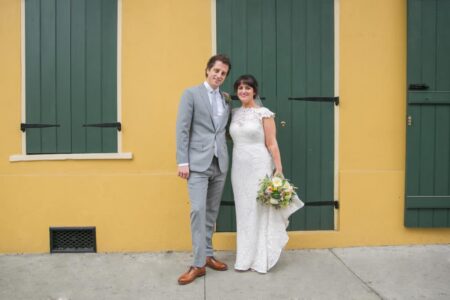 Smiling bride and groom in wedding attire pose by a yellow wall with green shutters; bride holds bouquet, groom in grey suit.