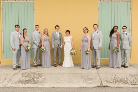 Bride, groom, bridesmaids in gray, and groomsmen in light gray pose smiling with flowers before a yellow wall with green doors.