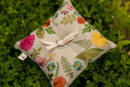 Colorful embroidered pillow with two silver rings tied by a cream ribbon, resting on green foliage.