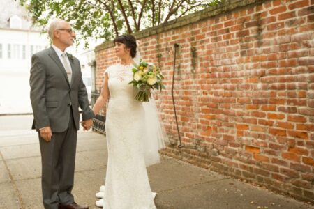 Bride in white dress holding bouquet, smiling with older man in gray suit by red brick wall and trees.