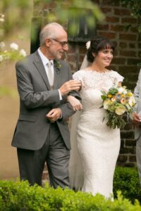 Bride in white lace dress and bouquet walks with smiling older man in gray suit near brick wall and greenery outdoors.