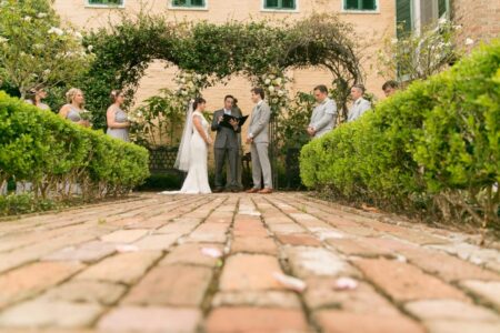 Bride and groom face each other outdoors at their wedding, with wedding party, greenery, and a brick path around them.
