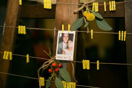 Smiling couple’s photo hangs on twine with yellow clothespins, green leaves, and red berries against a wood background.