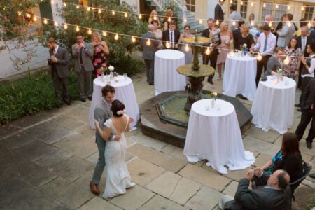 Bride and groom dance by a fountain at an outdoor wedding, with guests at round tables under string lights.