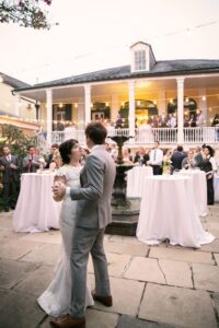 Bride and groom have their first dance outdoors, surrounded by guests, tables, string lights, and a two-story building.
