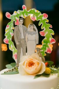 Bride, groom, and dog cake topper with floral decor on a white cake featuring a peach flower and greenery.