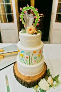 Three-tier white wedding cake with painted flowers, bride and groom topper, flower decoration, on a wooden slice stand.