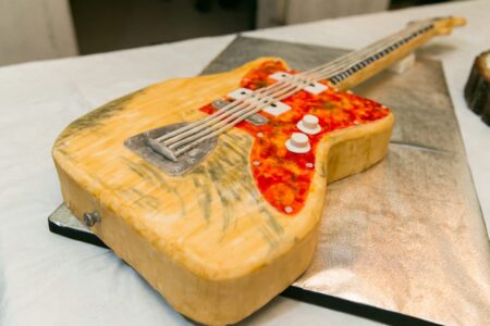 Electric guitar-shaped cake with strings, knobs, wood-grain details, displayed on a silver board.