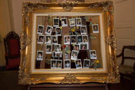 Gold ornate frame with Polaroid collage, flowers, and people, displayed on a wooden easel indoors.