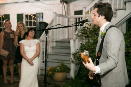 Man in suit plays guitar and sings at outdoor wedding as bride and smiling guests watch.