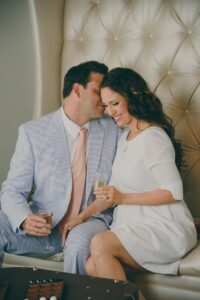 A formally dressed couple sits together on a plush chair, smiling and holding champagne glasses, looking happy and affectionate.
