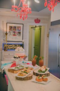 A stylish kitchen with a decorated table of appetizers, desserts, and drinks under a pink chandelier.