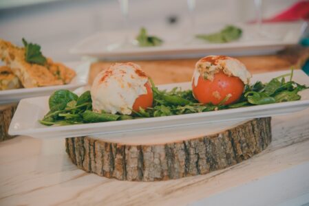 Stuffed tomatoes with melted cheese and herbs on fresh greens, served on a white plate atop a wooden slice.