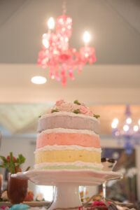 Pastel pink, yellow, and purple layer cake with decorative roses on a white stand; soft pink chandelier in background.