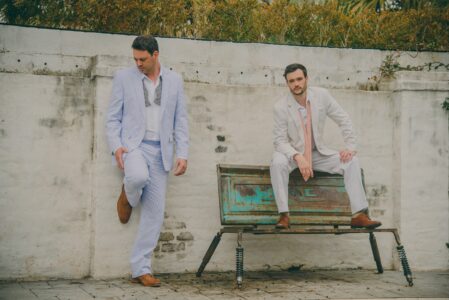 Two men in light suits pose outside—one stands, one sits on a bench—with brown shoes, greenery, and a white wall behind them.