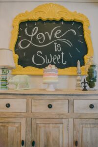 Rustic cabinet with pastel cake, knick-knacks, green pig figurine under a yellow-framed “Love is Sweet” chalkboard.