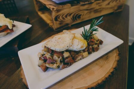 Hash of diced potatoes and vegetables with two fried eggs and rosemary on a white plate, served on a wooden slab.