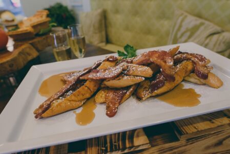 French toast with crispy bacon, powdered sugar, and syrup on a white plate; sparkling drinks and green seat in background.