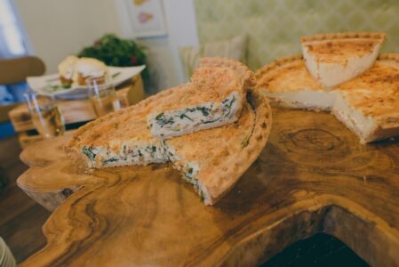 Golden quiche slices with creamy filling and spinach on a rustic platter; blurred table with more food and drinks in background.