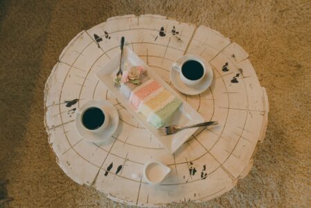 Overhead shot of two black coffees, cream, and pastel layered cake with forks on a rustic white table.