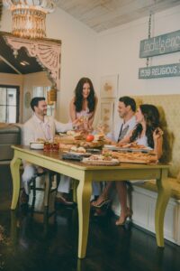 Four formally dressed adults smile and toast around a yellow dining table with food, in a stylish room with chandelier and art.
